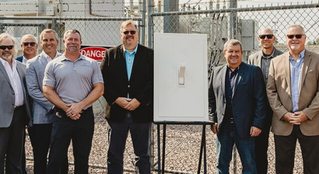 A photo of people standing next to a large light switch with a power substation in the background